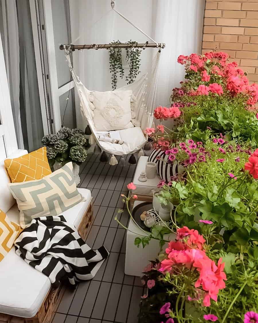 Cozy small balcony with a white hammock chair, soft beige cushions, and lush pink flowers in full bloom. A wooden pallet sofa with geometric-patterned pillows and a striped throw blanket adds to the inviting ambiance.