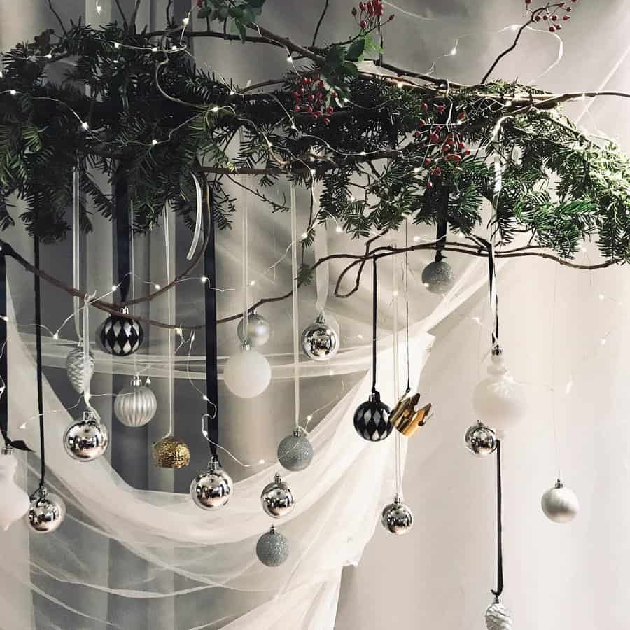 Hanging Christmas decorations with branches, string lights, and silver, white, and black ornaments against a sheer white background