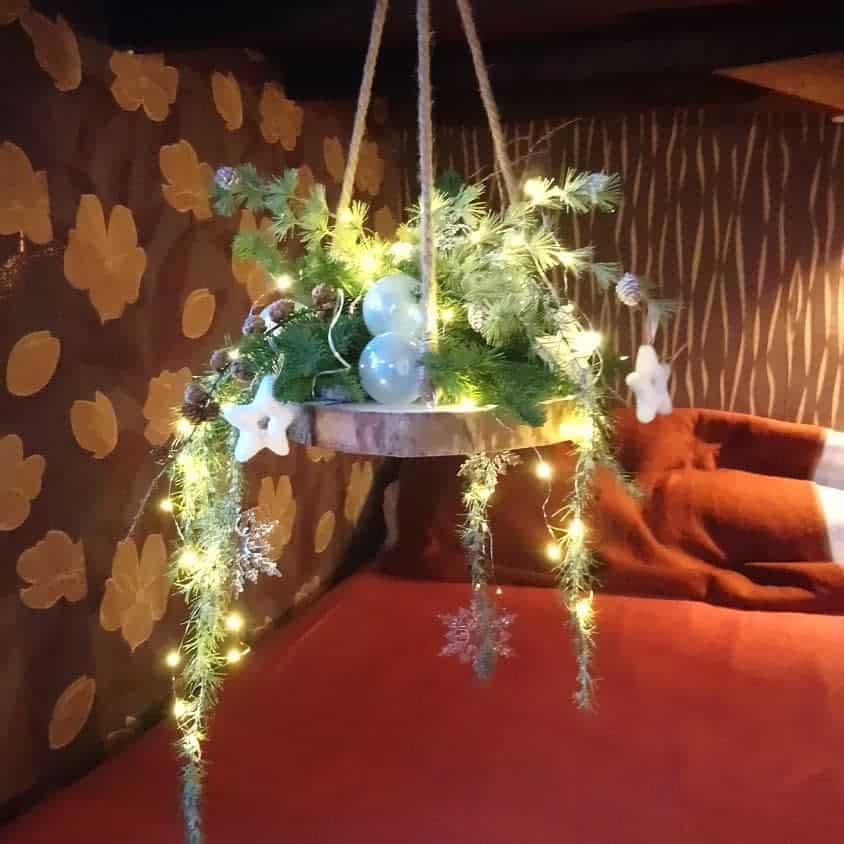 Hanging wooden wreath with greenery, fairy lights, and star ornaments, set against a floral patterned wall above a red bedspread