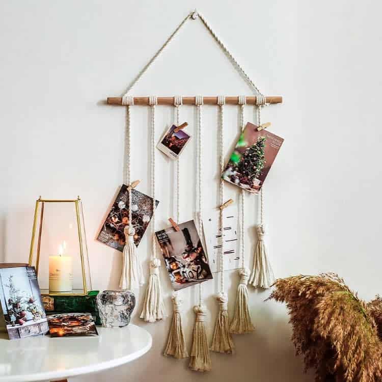 Wall decor with photos and tassels on strings, candle in glass holder, decorative vase, and dried plants on a nearby table