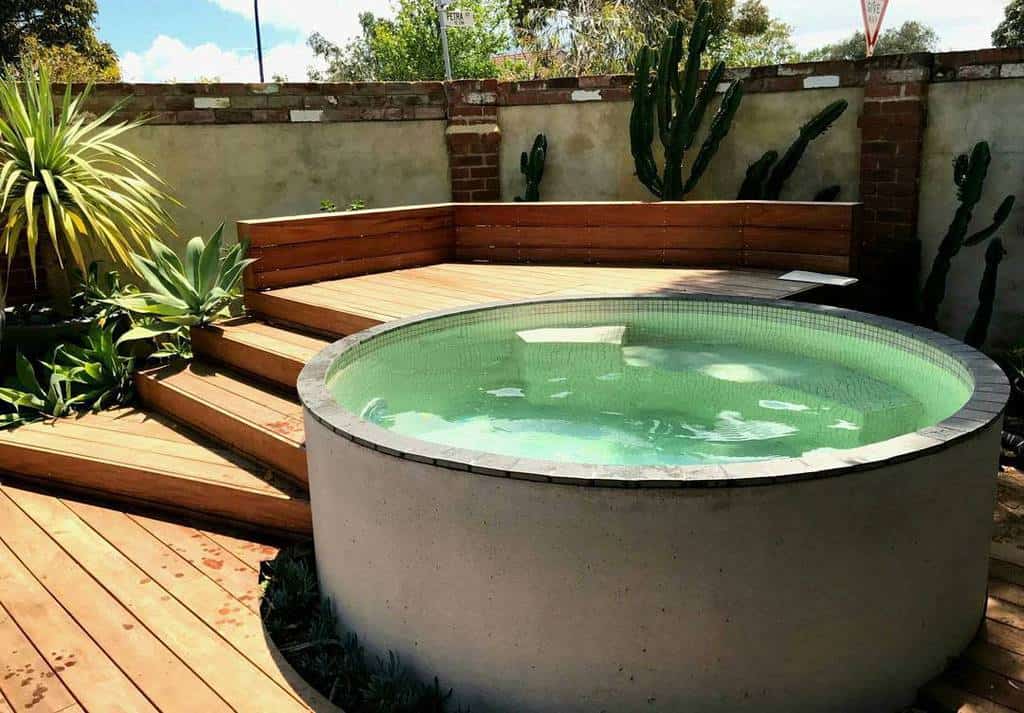 Stylish backyard with a circular plunge pool, wooden deck seating, lush succulents, and a brick privacy wall featuring cacti