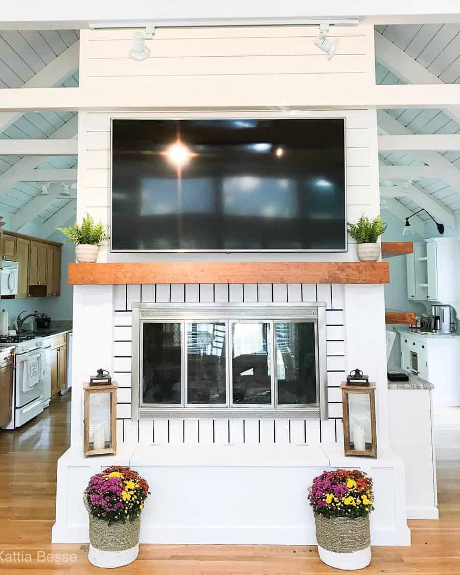 Living room with a TV above a fireplace, flanked by plants and lanterns; bright kitchen in the background with wooden floors