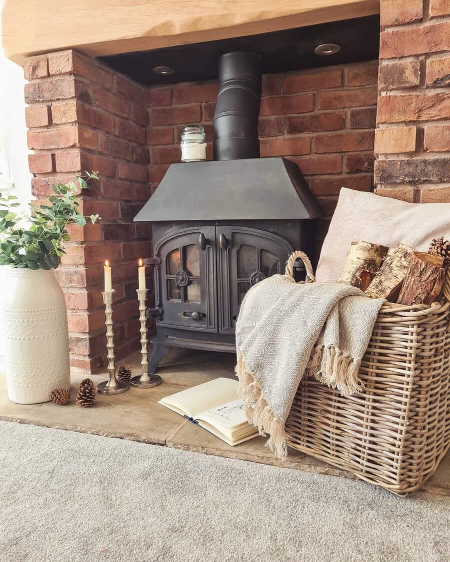 Cozy fireplace with a black stove, basket of firewood, candles, and an open book. Brick surround and a vase with greenery