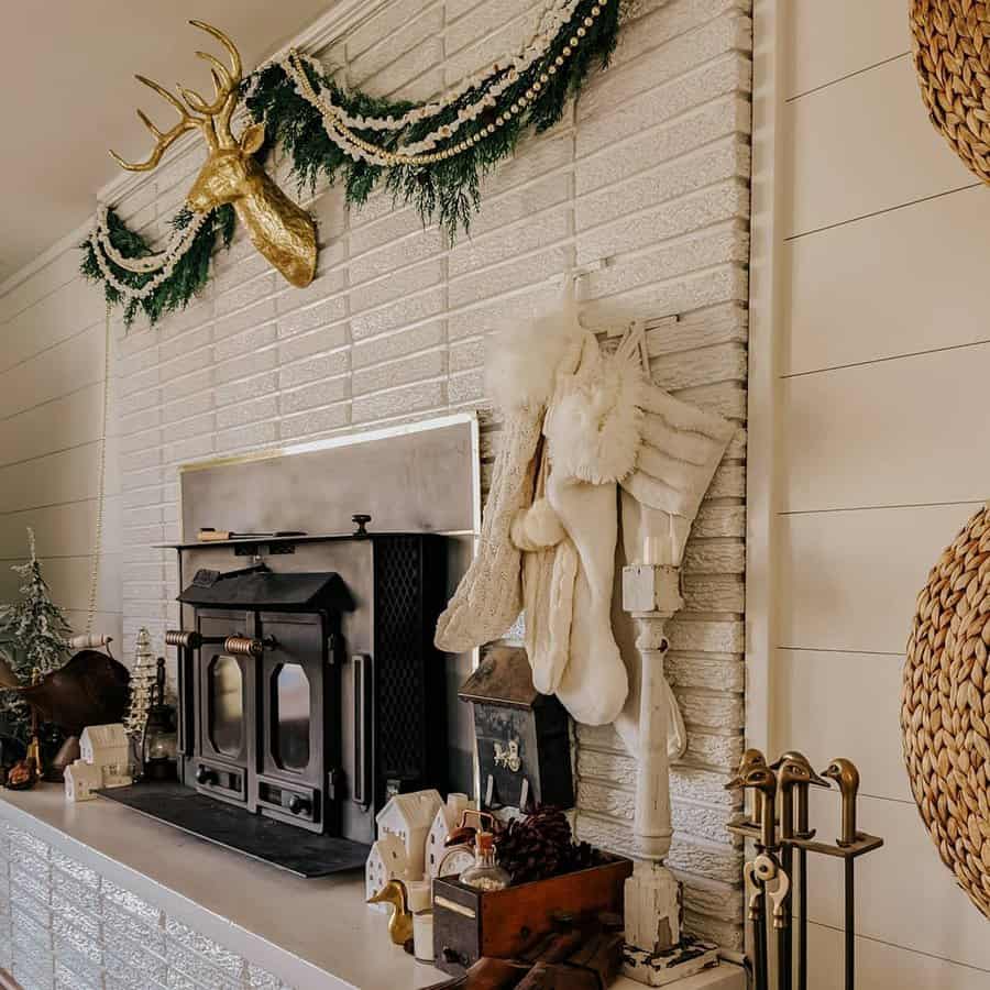 Cozy fireplace with stockings, festive decorations, and a gold deer head on a white brick wall. Wreaths and winter decor surround