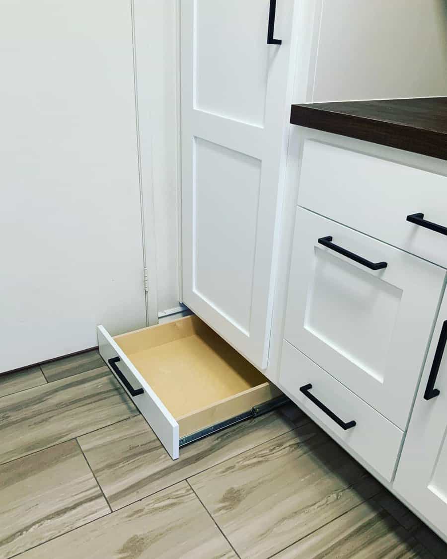 Open toe-kick drawer under white kitchen cabinets with black handles on a tiled floor