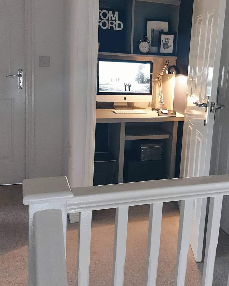 Compact home office in a closet with a desk, computer, lamp, and decorative items on shelves with door and banister in foreground