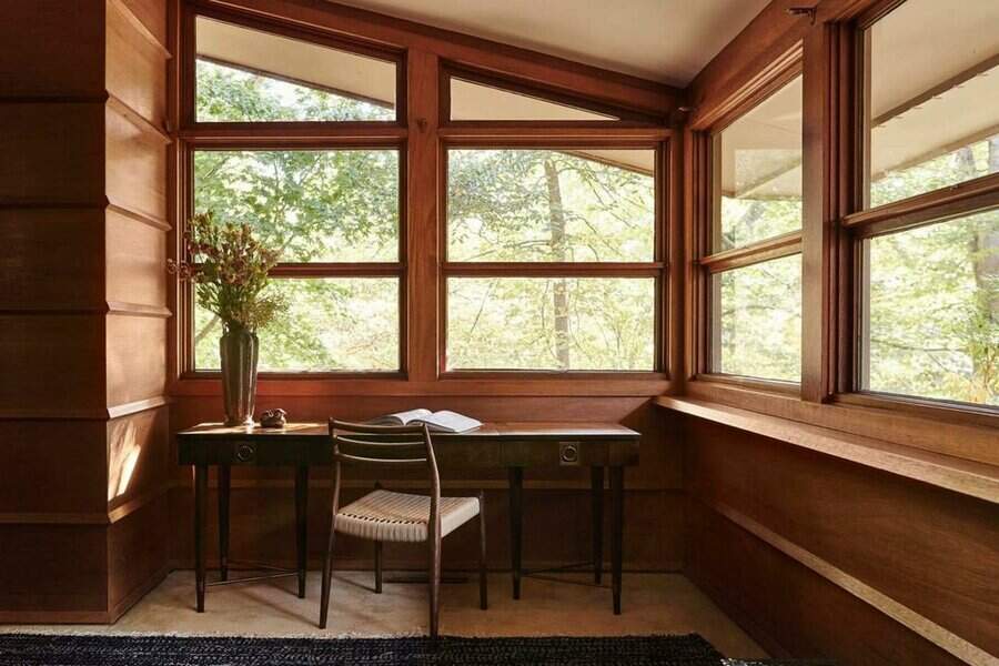 Cozy study nook with a wooden desk, chair, and large windows overlooking green trees, with a vase of flowers sitting on the desk