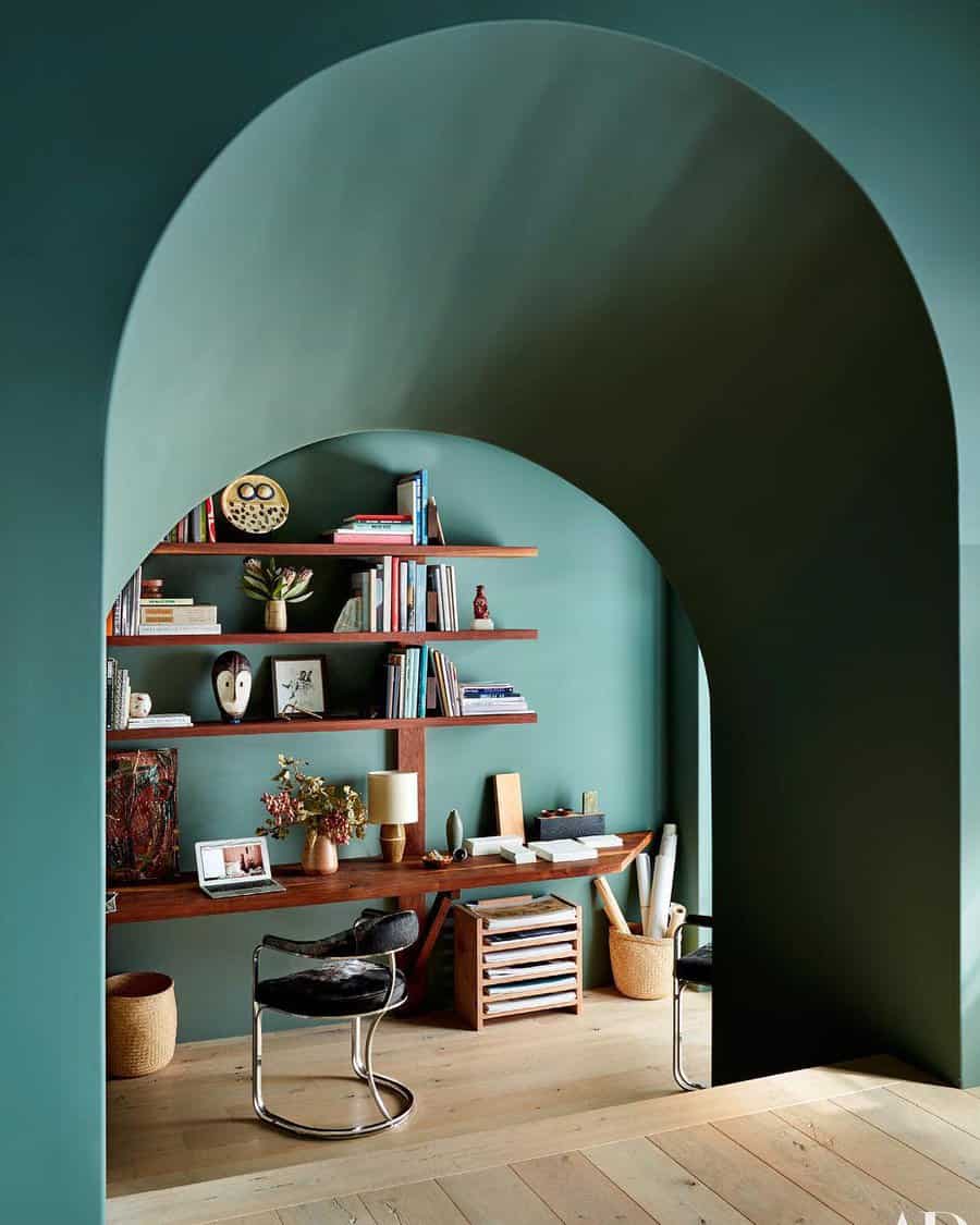 A cozy workspace with wooden shelves, books, decor, and a chair set within a teal-painted arched alcove