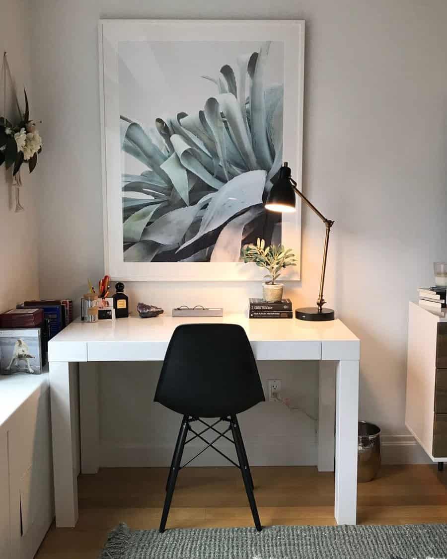 Minimalist desk setup featuring a black chair, desk lamp, books, and a plant with large plant artwork on the wall behind the desk