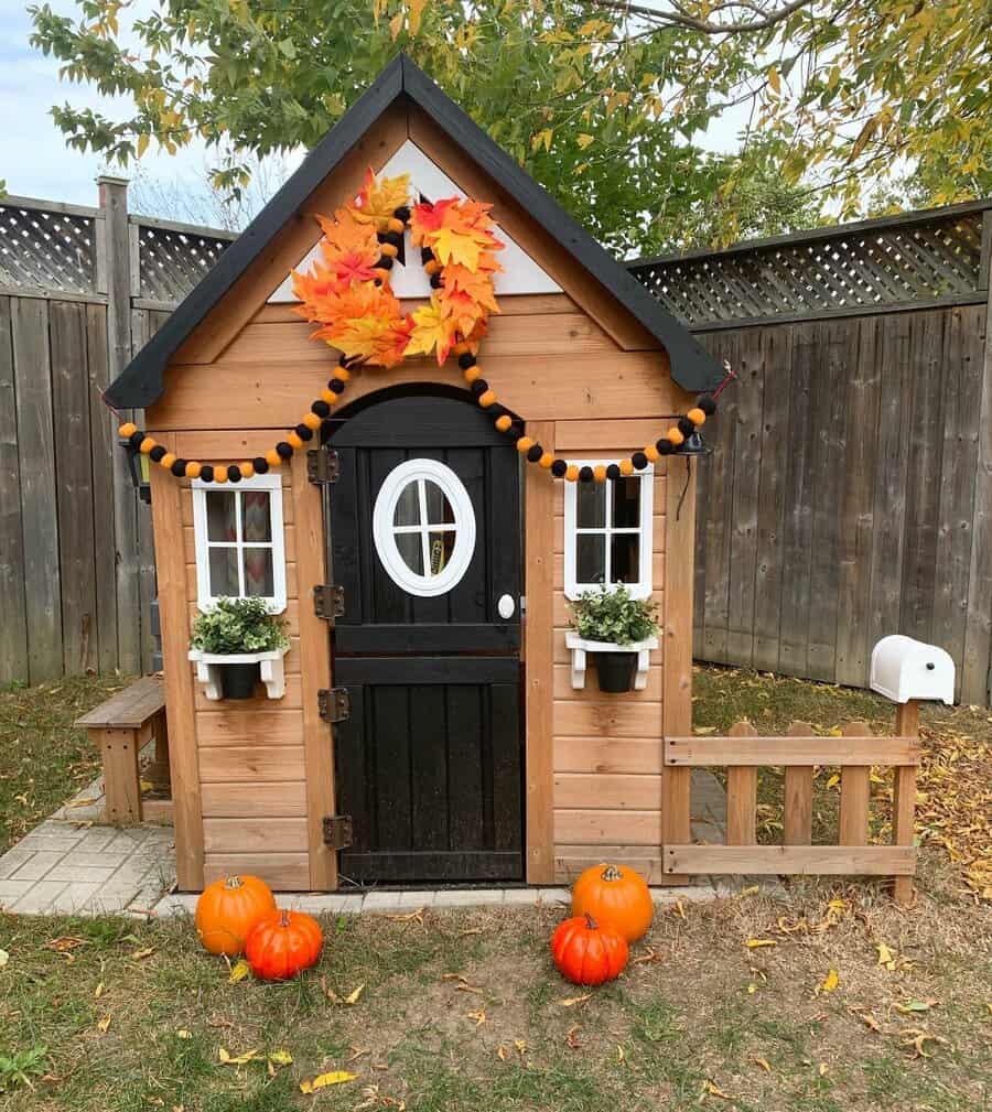 Small wooden playhouse decorated with orange and black garland, autumn leaves wreath, and pumpkins, surrounded by a wooden fence