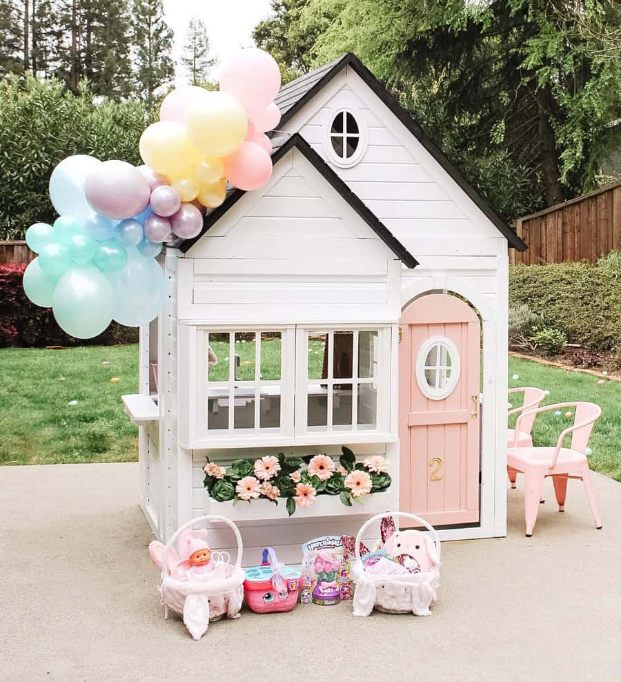 A white and pink playhouse with balloons, flowers, and toy baskets outside; two pink chairs sit nearby on a concrete patio