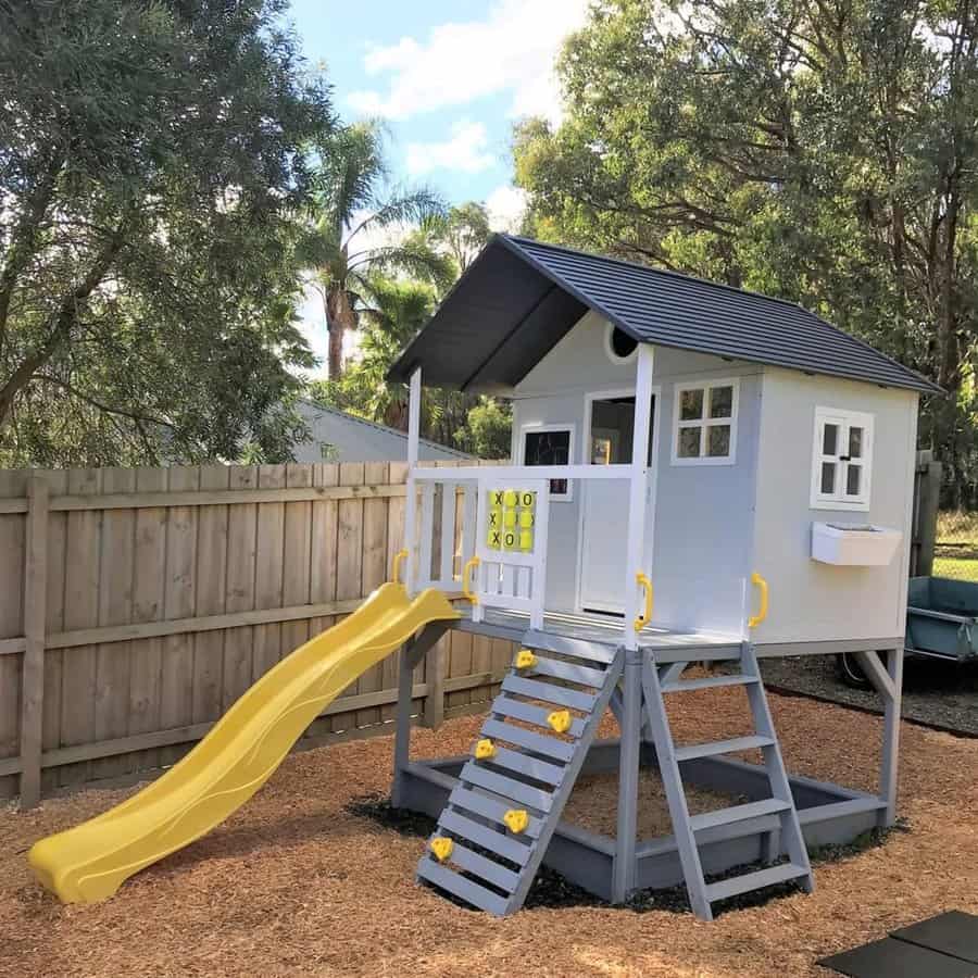 A grey and white playhouse on stilts with a yellow slide, ladder, and tic-tac-toe board, set in a wooden fenced yard with trees