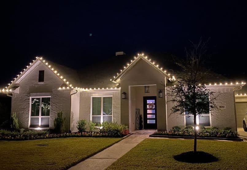 House with white brick exterior adorned with string lights along roofline, glowing warmly at night, surrounded by a trimmed lawn