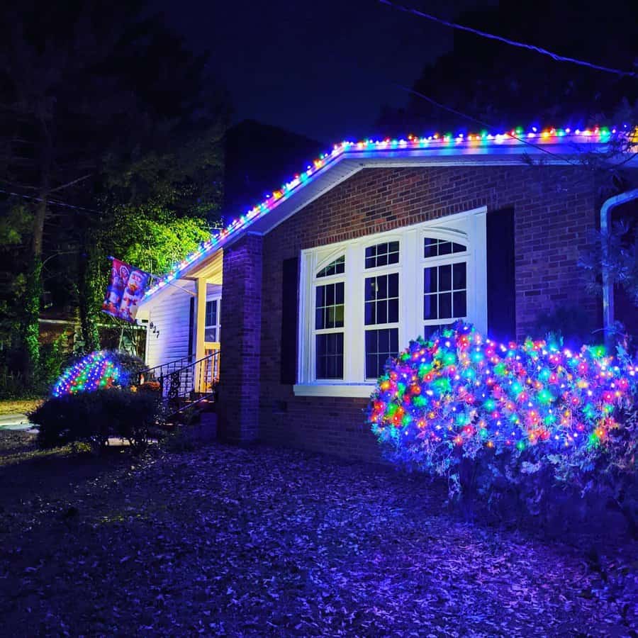 House with colorful lights decorating roofline and bushes, glowing at night; white-framed windows and brick facade visible