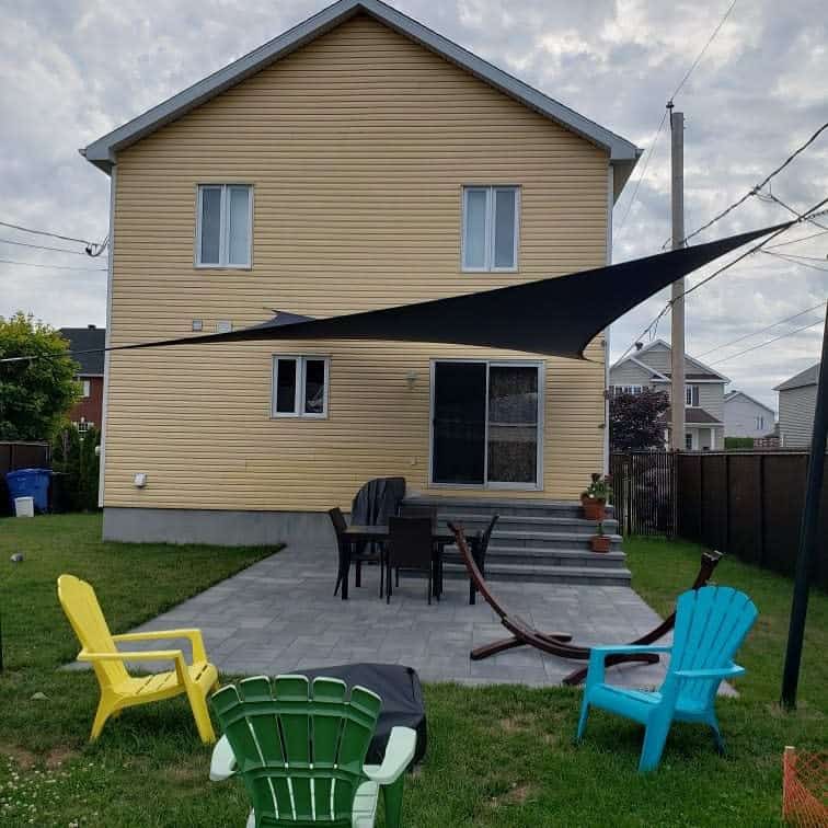 Backyard patio with a black shade sail, outdoor dining set, colorful Adirondack chairs, and a hammock near a yellow house