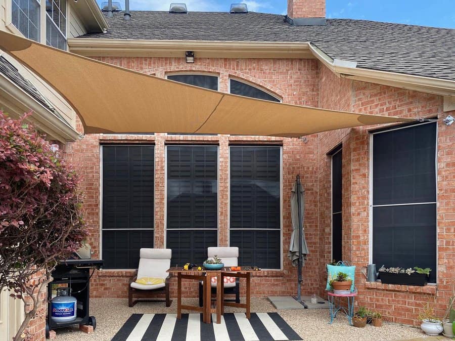 Brick patio with a tan shade sail, lounge chairs, a striped rug, and potted plants, creating a cozy outdoor seating area