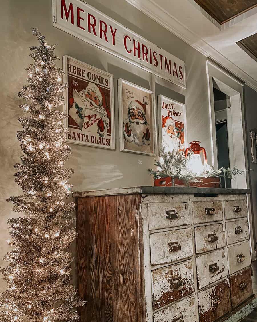 Vintage Christmas decor with a small lit tree, "Merry Christmas" sign, and retro Santa artwork above a rustic wooden dresser