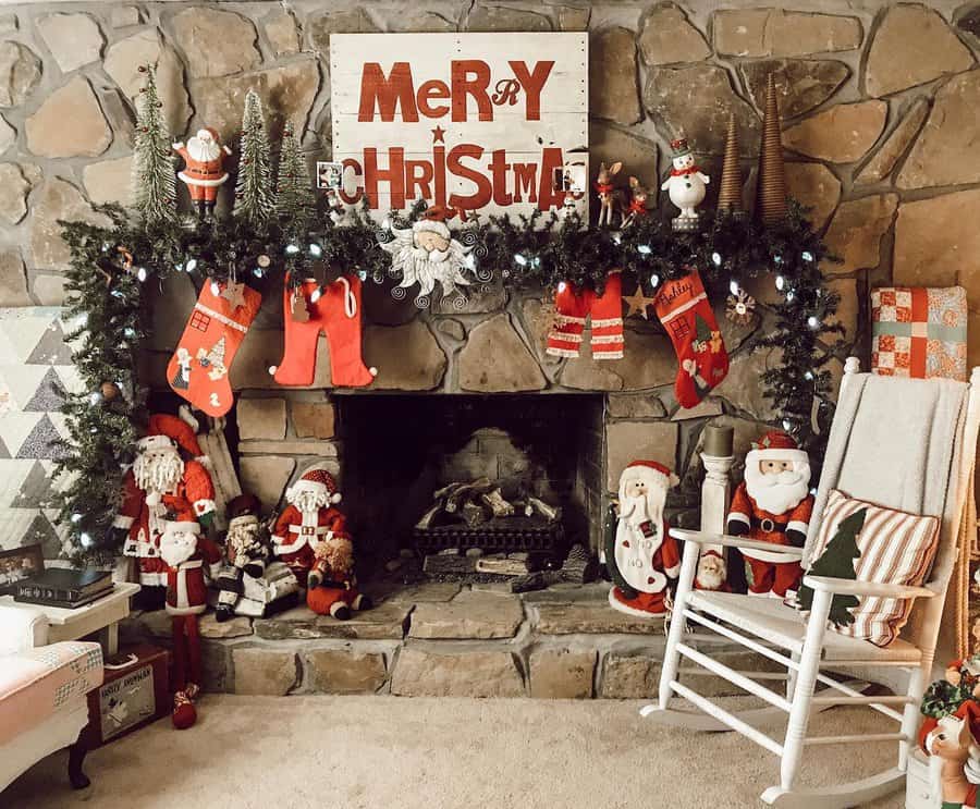 A cozy fireplace decorated with stockings, Santa figures, and a "Merry Christmas" sign on a stone wall, with a rocking chair on the side