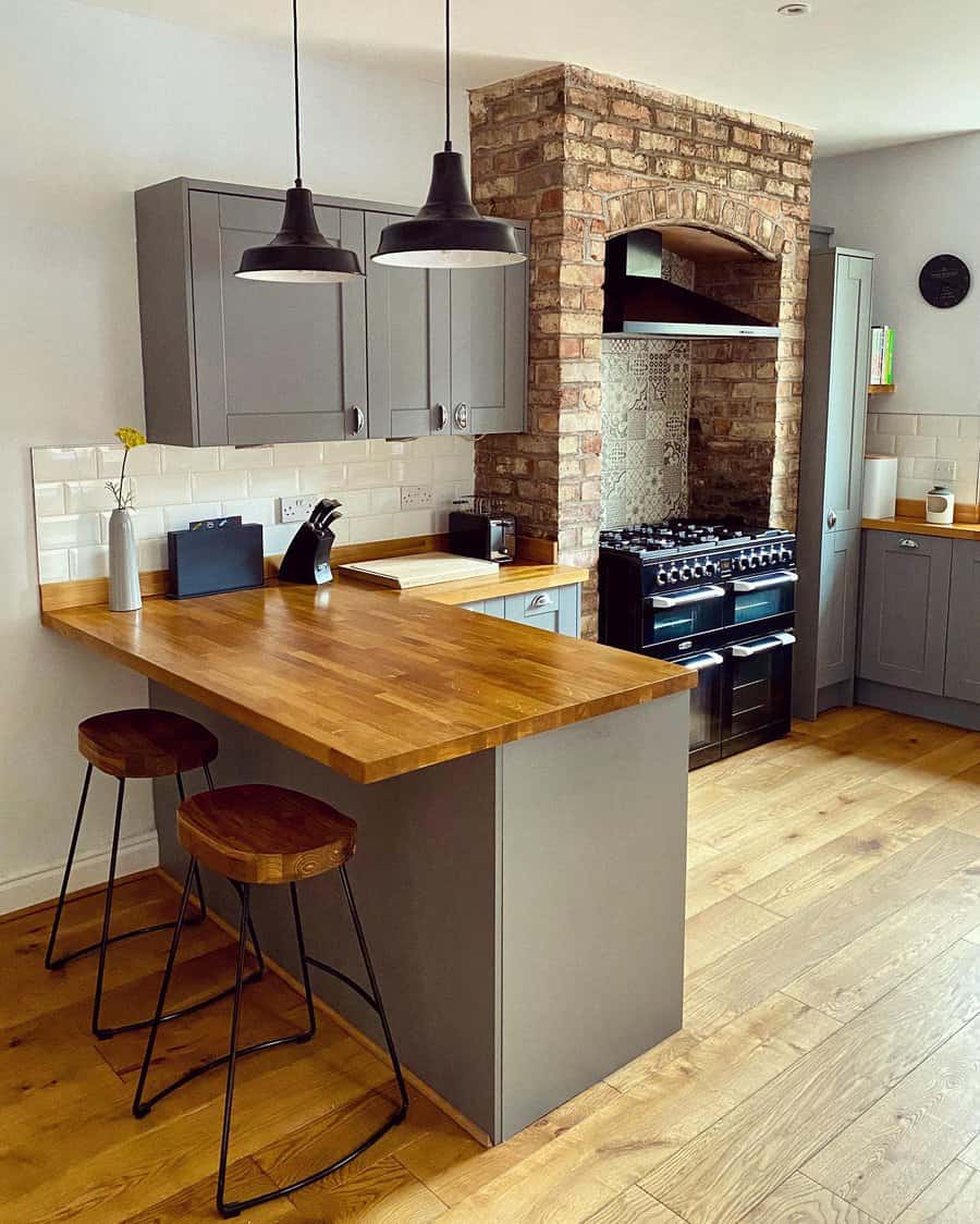 Industrial-style kitchen with a wooden breakfast bar, gray cabinetry, exposed brick feature, black pendant lighting, and rustic barstools