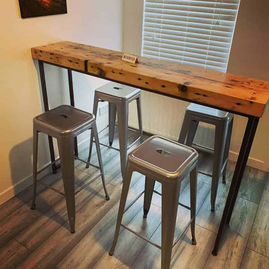 A rustic wooden high-top table with four gray metal stools in a room with light wood flooring and blinds on the window