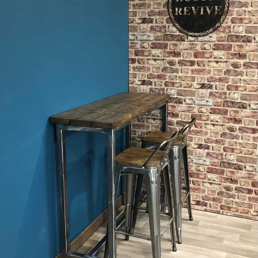 Wooden high table with two metal bar stools against a blue and brick-patterned wall, with a partially visible round sign above