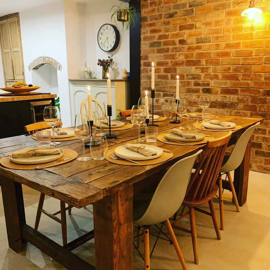 A rustic dining table set for six with elegant place settings, candles, and a warm brick wall backdrop