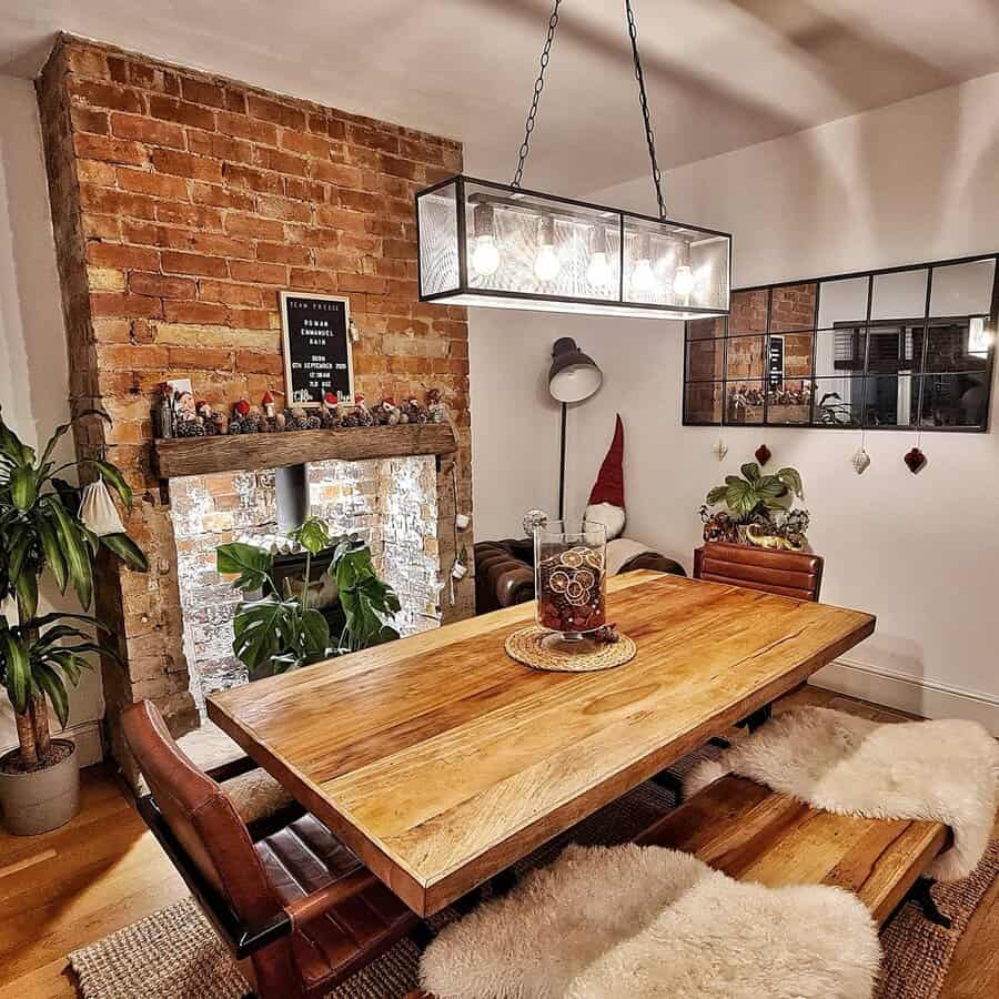 Cozy dining area with a wooden table, leather chairs, brick accent wall, and a hanging light fixture, decorated with plants and a mirror