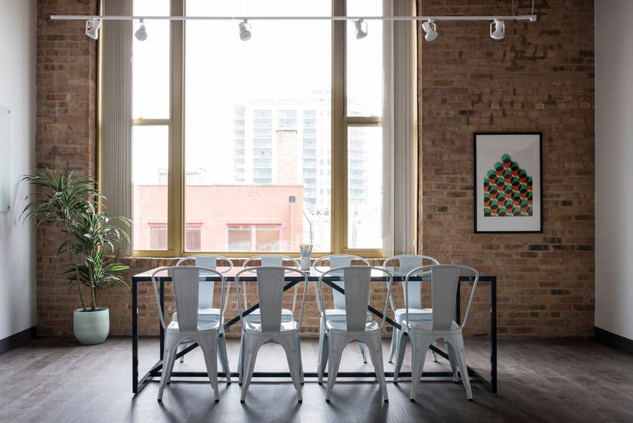 Bright dining room with brick walls, large windows, a metal table with eight chairs, plant, and framed artwork on the wall