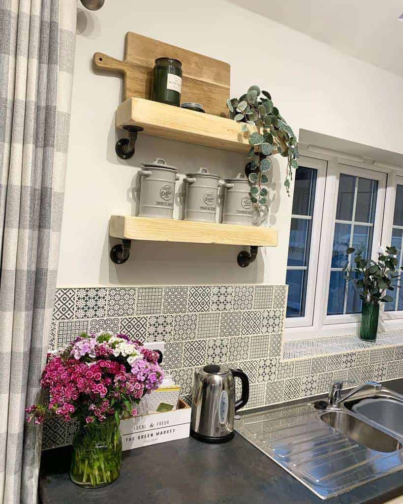 Kitchen corner with patterned tiles, wooden shelves holding jars and plants, a bouquet of flowers, kettle, and sink by a window