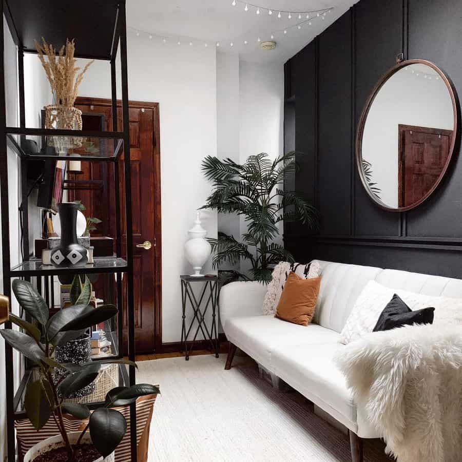 Tiny living room with a white sofa, various pillows, a large mirror, tall plants, and a bookshelf against a black accent wall