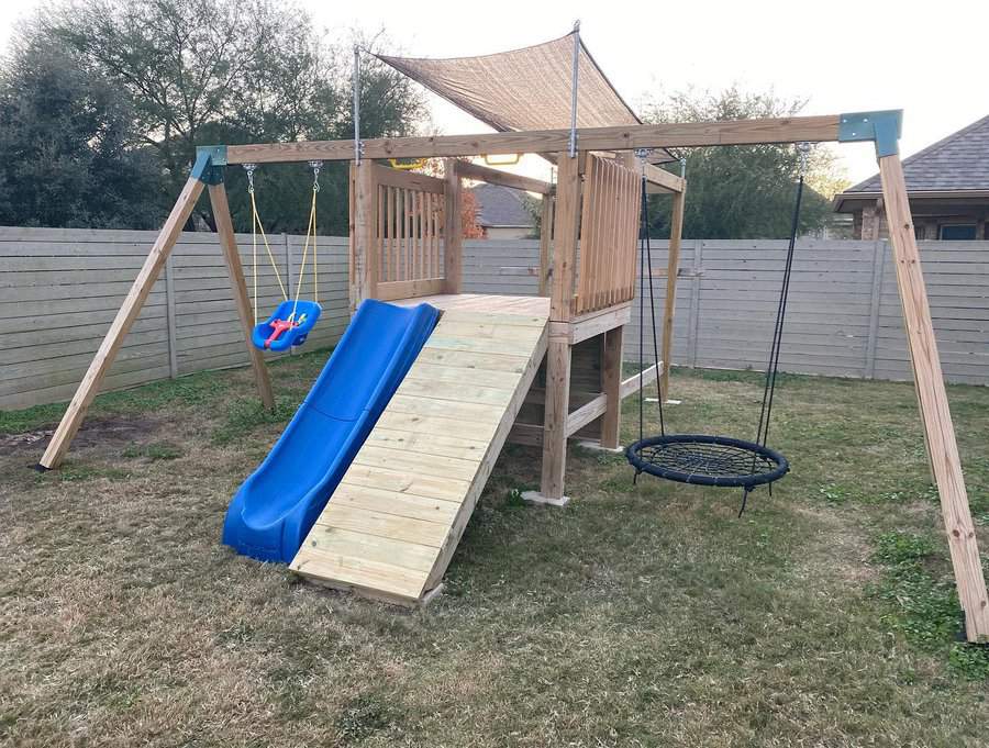 Wooden playground set with slide, swings, and a rope swing, surrounded by a fenced yard with grass and trees in the background