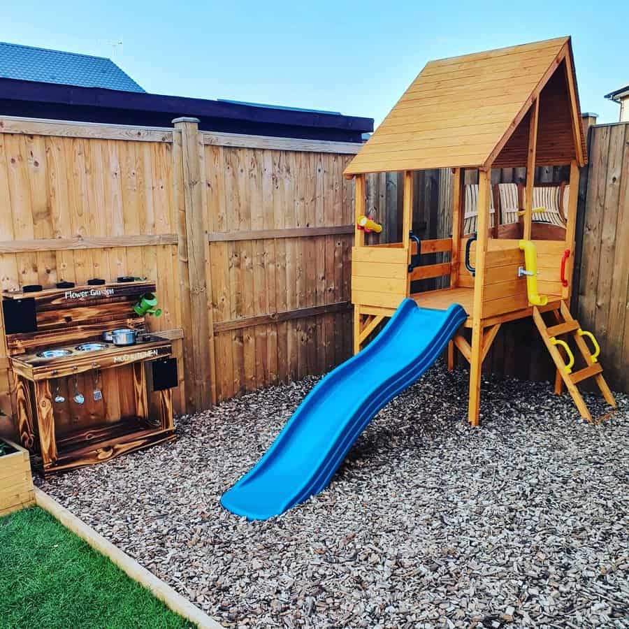 Compact wooden backyard playground with a blue slide, climbing ladder, and playhouse, next to a wooden mud kitchen for creative play