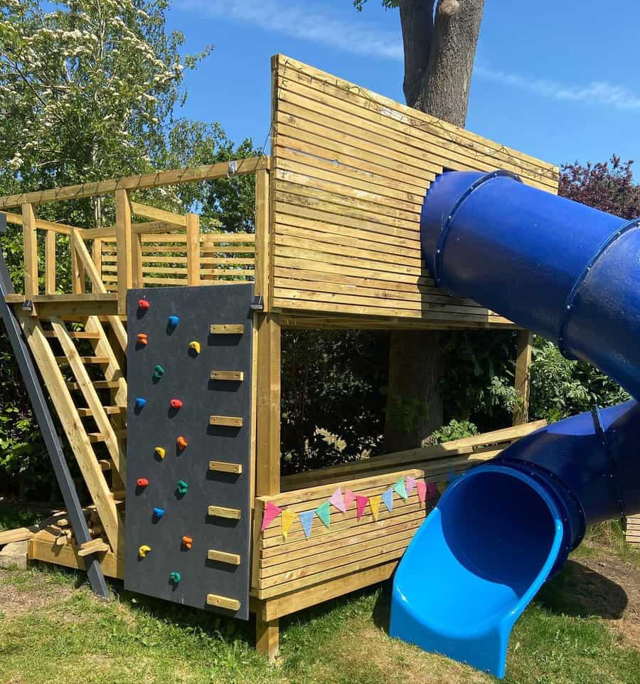 Wooden play structure with a climbing wall and a blue spiral slide, decorated with colorful bunting, set in a garden