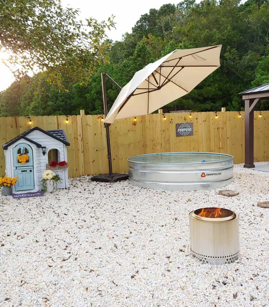 Backyard with a doghouse, large umbrella, metal pool, and a fire pit on a gravel surface, surrounded by a wood fence