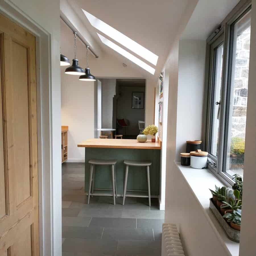 Cozy breakfast bar with a wooden countertop, sage green base, minimalist barstools, skylights, and industrial pendant lighting for a warm ambiance