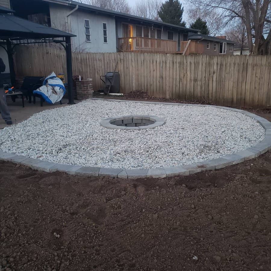 Backyard fire pit area with circular gravel layout, stone edge, and a central pit; wooden fence and house in the background