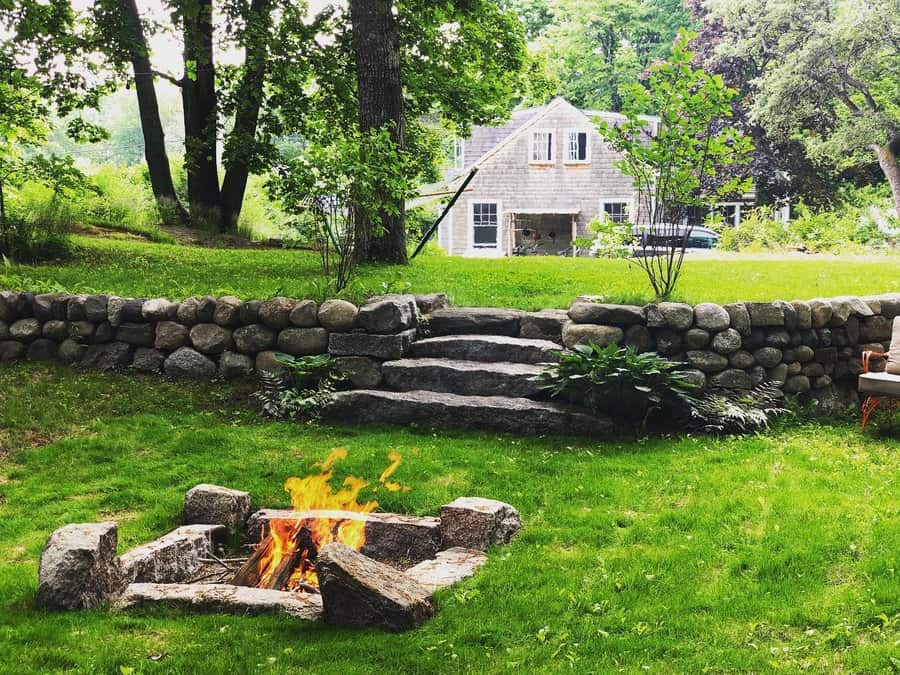 Backyard scene with a stone fire pit, green lawn, stone steps, and a cottage in the background surrounded by trees