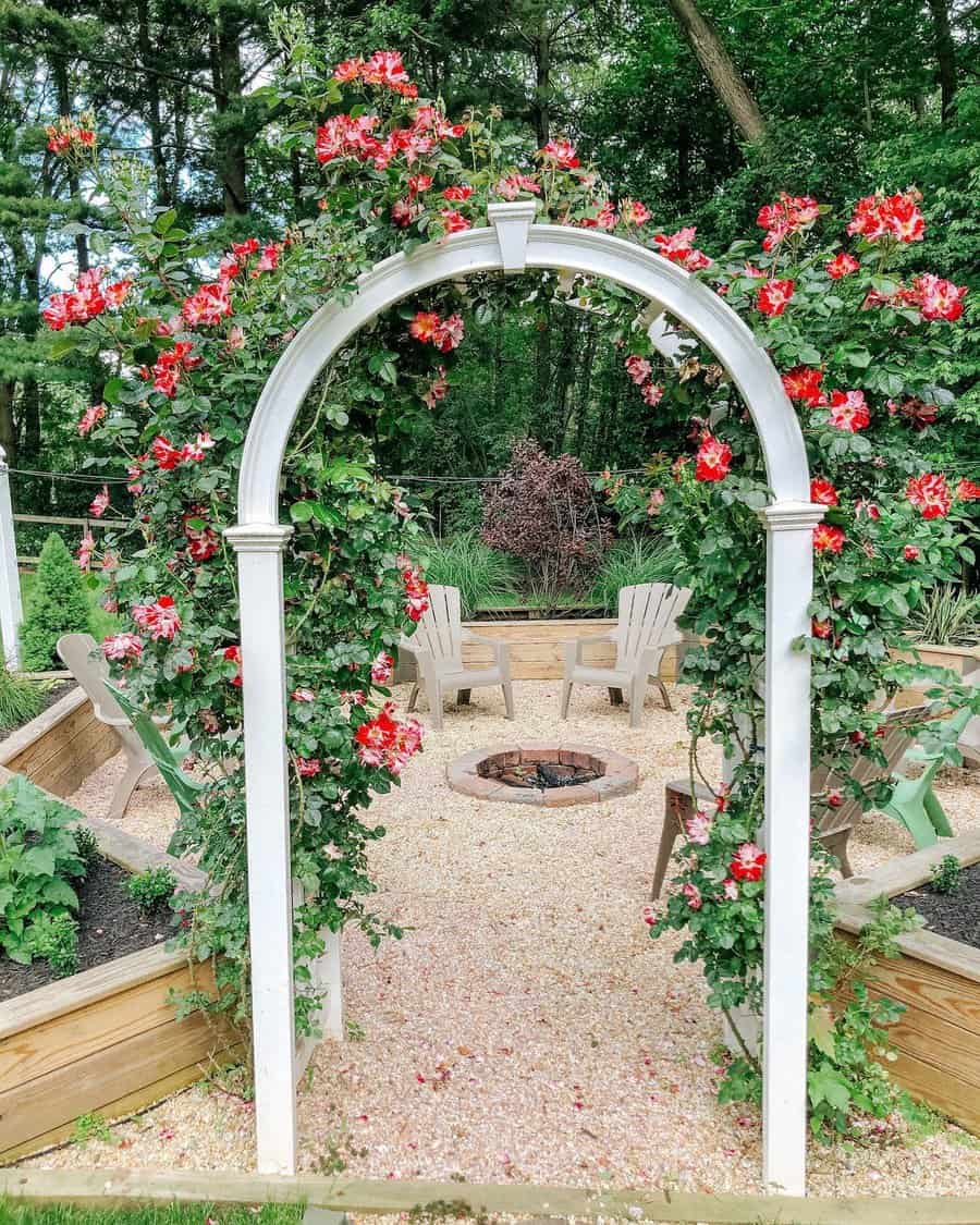 White arch with red roses leads to a gravel seating area with Adirondack chairs and a fire pit, surrounded by lush greenery