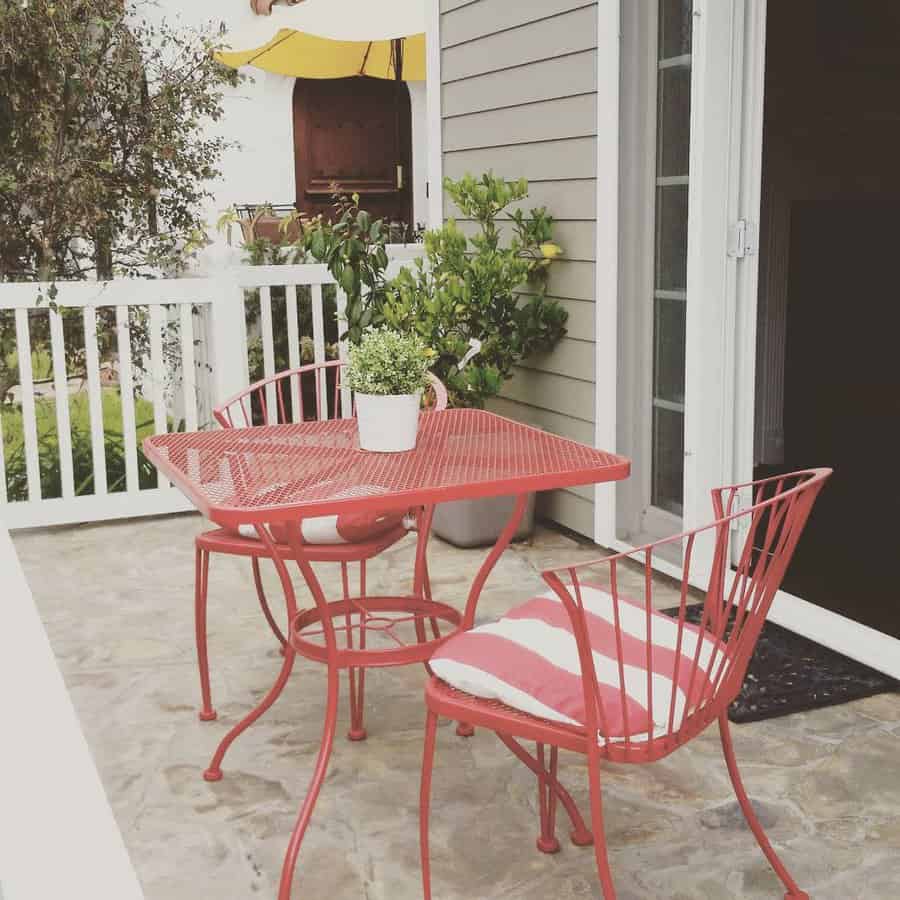 Red patio set with a table and two chairs, one with a striped cushion. Potted plant centerpiece. White fence and door in the background