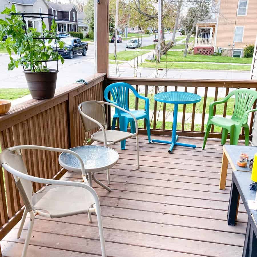 Wooden porch with assorted chairs around two small tables. A potted plant is on the railing, and houses are visible in the background