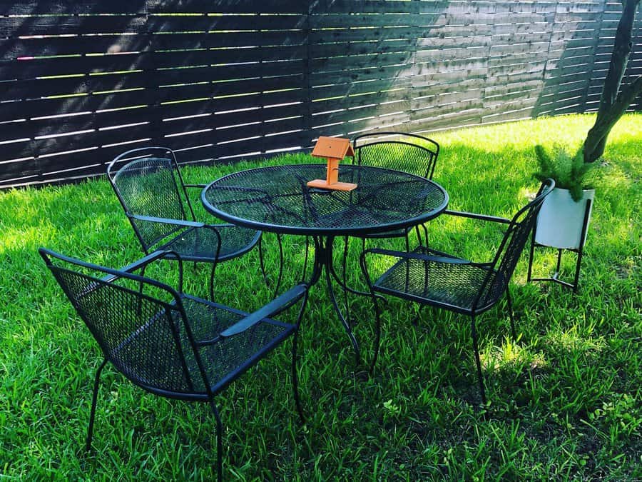 Black metal patio set with a round table and four chairs on grass, featuring a small wooden birdhouse centerpiece and a wooden fence in the background