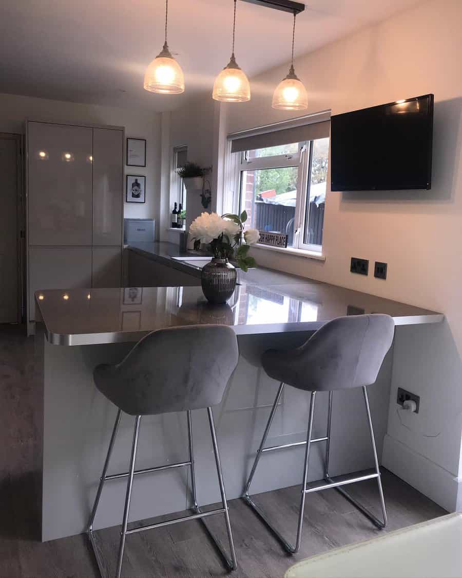 Modern kitchen with a sleek gray breakfast bar, plush velvet barstools, glass pendant lighting, and a mounted TV for a cozy and stylish look