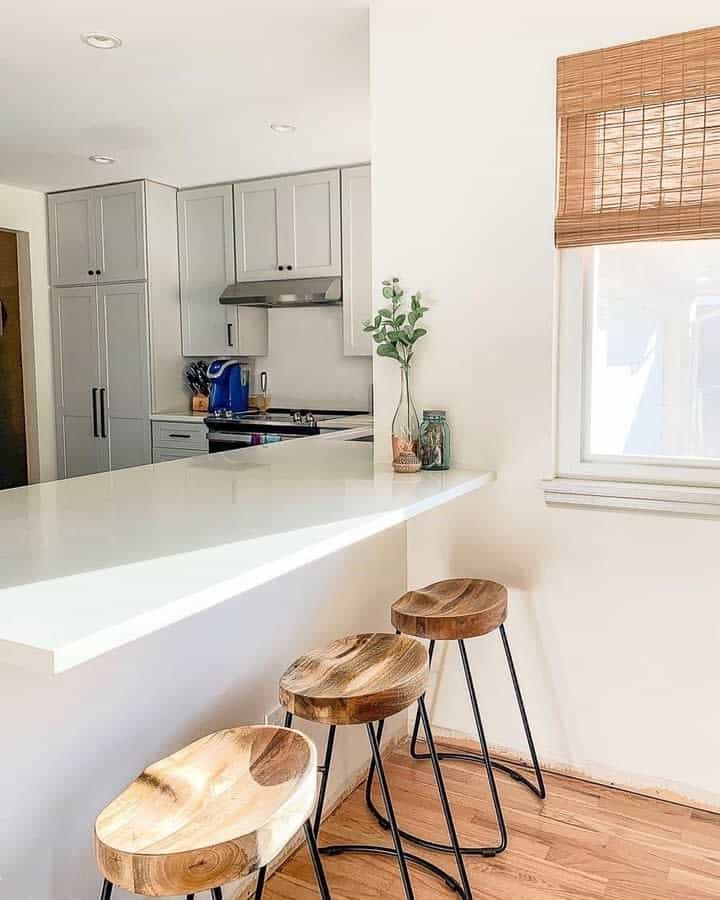 Bright kitchen with a white quartz breakfast bar, wooden and metal barstools, light gray cabinetry, and natural woven blinds for a cozy touch