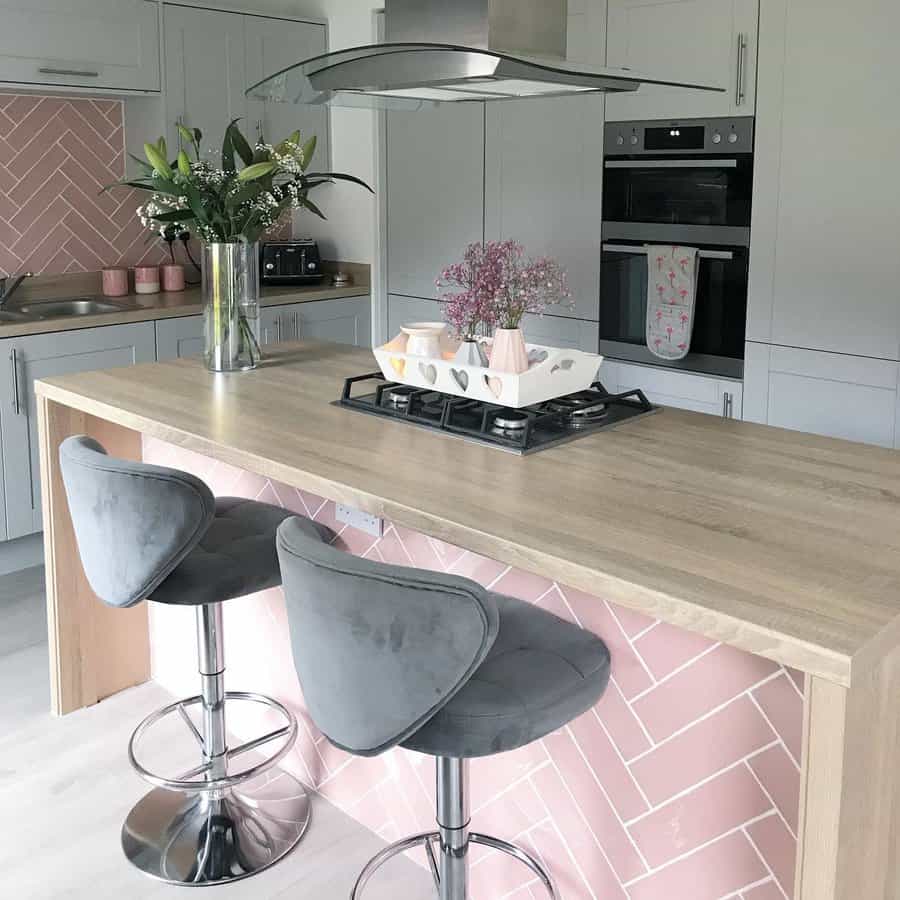 Chic kitchen with a wooden breakfast bar, pink herringbone tile accent, plush gray barstools, and soft pastel decor for a cozy, modern look