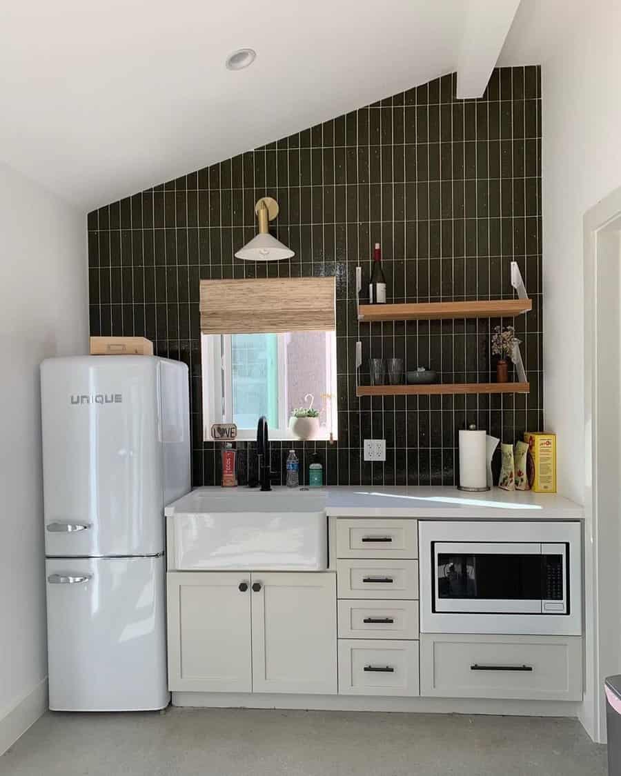 Small kitchen with a white fridge, sink, microwave, green tiled backsplash, shelves with bottles, and a small window