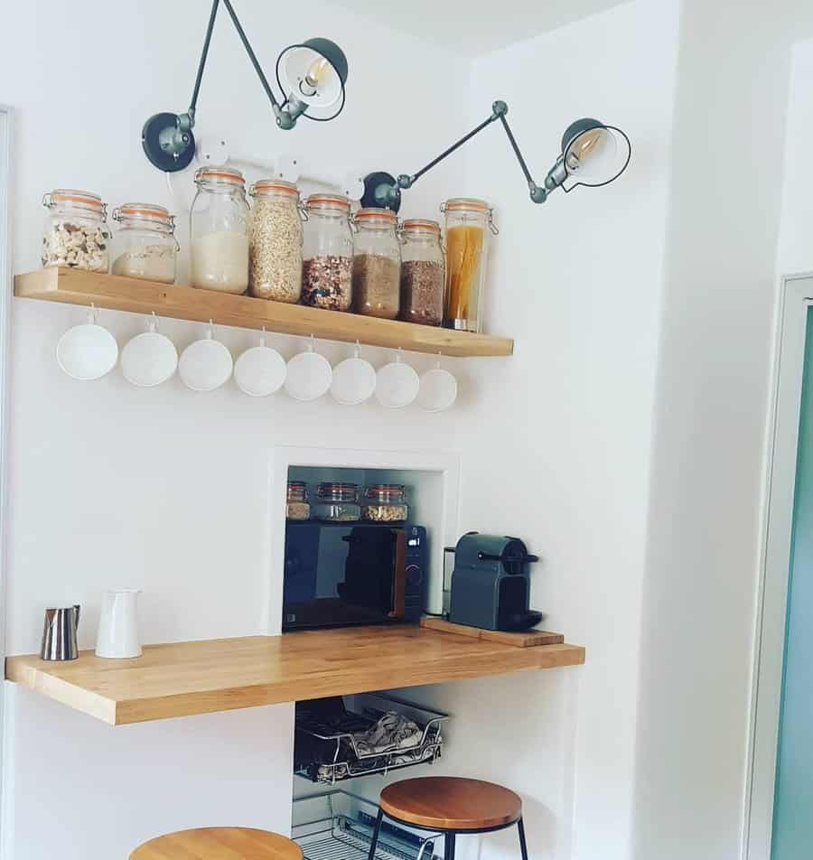 Minimalist kitchen corner with wooden shelves, jars of grains, hanging cups, a coffee machine, and wall-mounted lamps