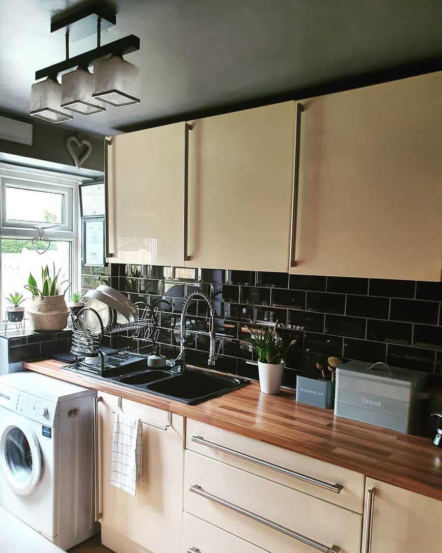 Modern kitchen with beige cabinets, black tile backsplash, wooden countertops, plants, a washing machine, and a sink with dishes