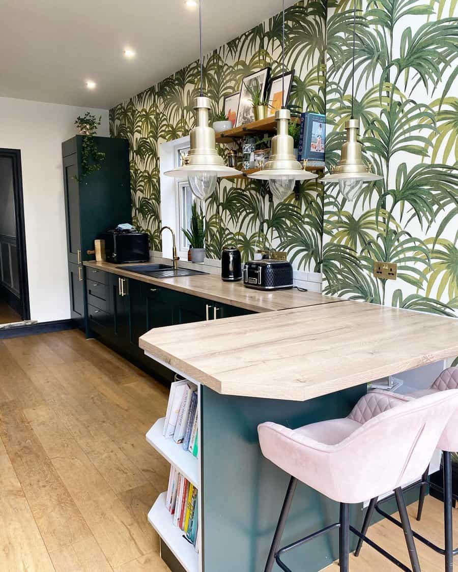 Kitchen with tropical leaf wallpaper, wooden countertops, green cabinets, pendant lights, and barstools. Shelves hold decor and cookbooks