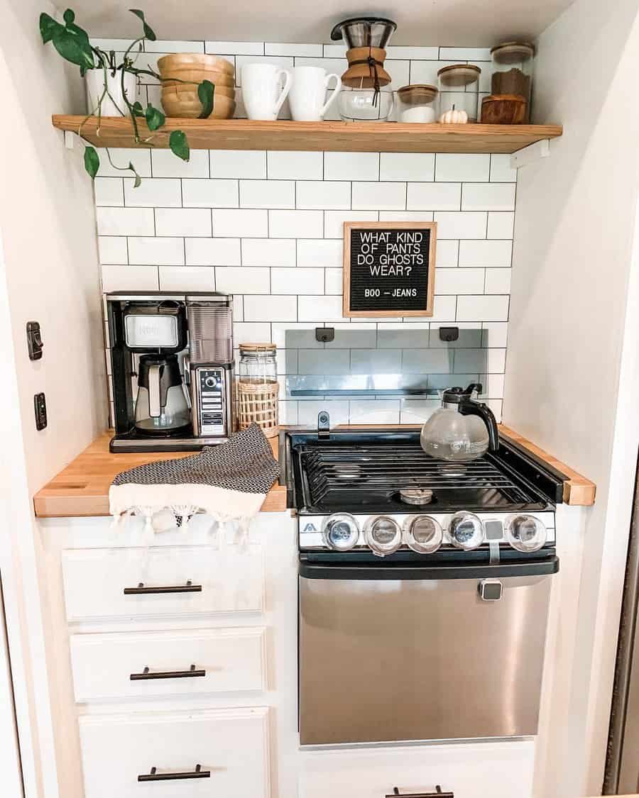 A cozy RV kitchenette with white tiles, a coffee maker, a stovetop, and a fun Halloween-themed sign, creating a welcoming and functional space.