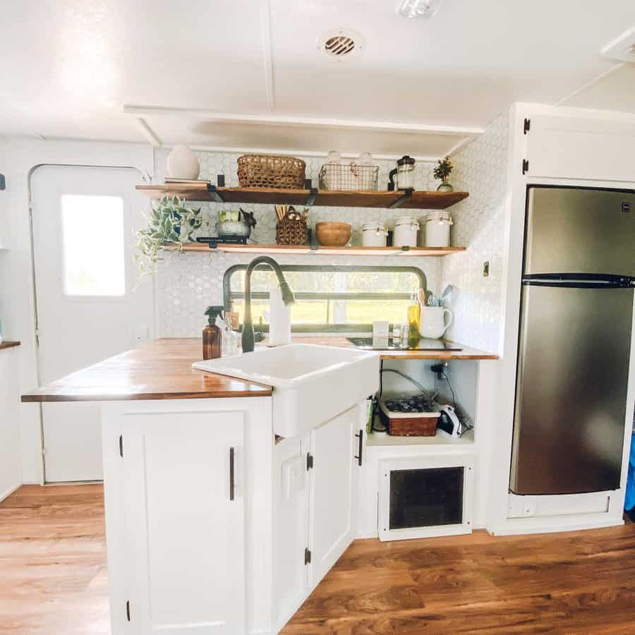 A clean and modern RV kitchenette with wooden shelves, a large sink, and neatly arranged plants and kitchen essentials, offering a bright and functional space.